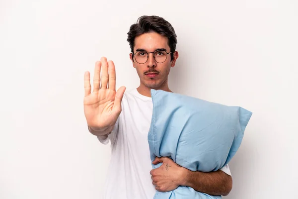 Young caucasian man wearing pajamas holding a pillow isolated on white background standing with outstretched hand showing stop sign, preventing you.