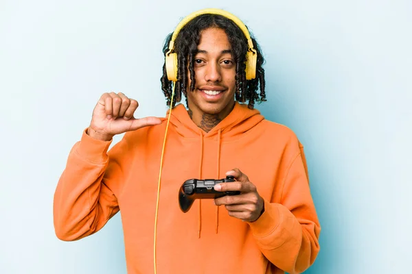 Young African American man playing with a video game controller isolated on blue background feels proud and self confident, example to follow.