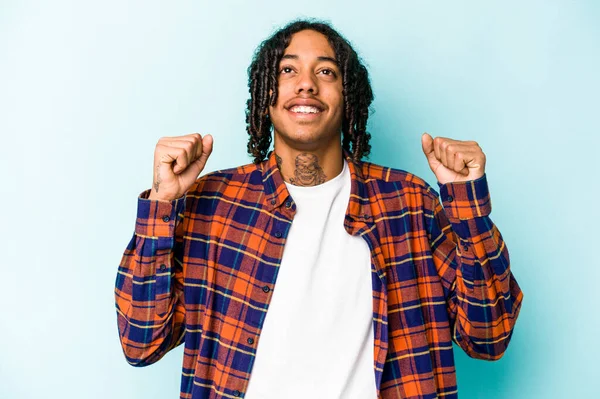 Genç Afrikalı Amerikalı bir adam mavi arka planda izole edilmiş zafer, tutku ve coşkuyu kutluyor, mutlu bir ifade..