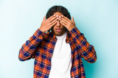 Genç Afrikalı Amerikalı bir adam mavi arka planda izole edilmiş. Gözleri elleriyle kapatmaktan korkuyor..