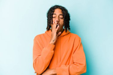 Genç Afrikalı Amerikalı bir adam mavi arka planda izole edilmiş esniyor ve ağzı eliyle kapalı yorgun bir jest yapıyor..