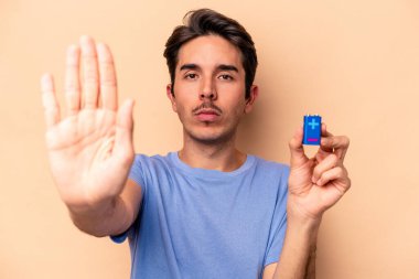 Beyazlı genç bir adam bej arka planda izole edilmiş bir bateriyi tutuyor. Elini uzatıp dur işareti veriyor..