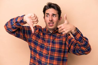 Beyaz tenli genç adam bej arka planda izole edildi. Baş parmağını kaldırdı ve baş parmağını kaldırdı. Zor seçim kavramı.