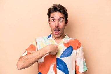 Beyaz tenli genç adam bej arka planda izole edildi. Parmakla göstererek şaşırdı, gülümsedi..