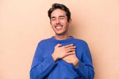 Beyazlı genç bir adam bej arka planda izole edilmiş. Gülüyor, elini kalbe koyuyor, mutluluk kavramını koruyor..