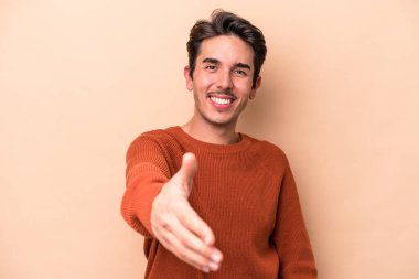 Beyazlı genç bir adam bej arka planda izole edilmiş. Kameraya doğru elini uzatarak selam veriyor..