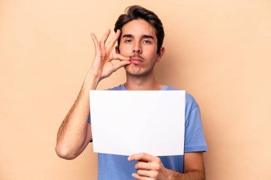 Beyazlı genç bir adam bej arka planda izole edilmiş bir pankart tutuyor ve parmakları dudaklarında bir sır saklıyor..