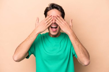 Beyaz tenli, bej arka planda izole edilmiş genç bir adam elleriyle gözlerini kapatıyor, gülümseyerek büyük bir sürpriz bekliyor..