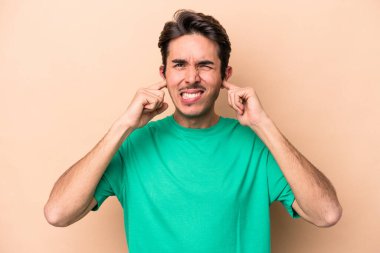 Beyazlı genç bir adam bej arka planda izole edilmiş parmaklarıyla kulaklarını kapatıyor, gürültülü bir ortam yüzünden stresli ve çaresiz..