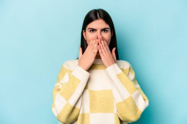 Mavi arka planda izole edilmiş genç beyaz kadın şok olmuş, ağzı ellerle kaplanmış, yeni bir şey keşfetmeye hevesli..