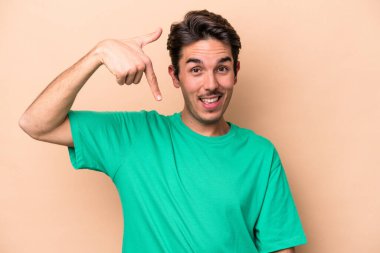 Beyazlı genç bir adam bej arka planda izole edilmiş. Elle bir gömlek fotokopi alanını işaret ediyor. Gururlu ve kendinden emin.