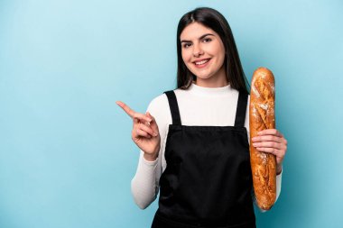 Genç fırıncı Kafkas elinde mavi arka planda izole edilmiş bir somun ekmek tutuyor gülümsüyor ve boş alanda bir şey gösteriyor..