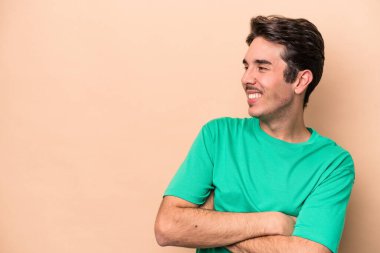 Beyaz tenli genç adam bej arka planda izole edilmiş. Kollarını kavuşturup gülümsüyor..