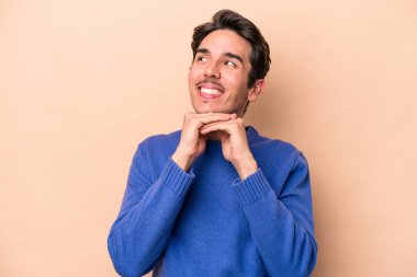 Beyaz tenli genç adam bej arka planda izole edilmiş ellerini çenesinin altında tutuyor, mutlu bir şekilde yana bakıyor..