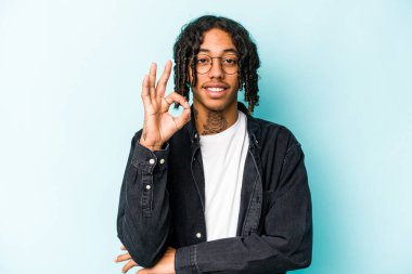 Genç Afrikalı Amerikalı bir adam mavi arka planda izole edilmiş bir şekilde bir göz kırpıyor ve elleriyle iyi bir jest yapıyor..