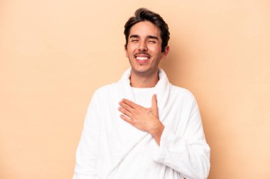 Kafkasyalı genç adam bej arka planda izole edilmiş bornoz giyiyor kahkahalar atıyor göğsünde elini tutuyor.