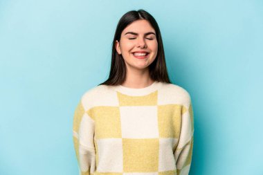 Mavi arka planda izole edilmiş genç beyaz kadın gülüyor ve gözlerini kapıyor, rahatlamış ve mutlu hissediyor..