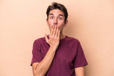 Beyaz tenli genç adam bej arka planda izole edilmiş şok olmuş, ağzı ellerle kaplanmış, yeni bir şey keşfetmeye hevesli..