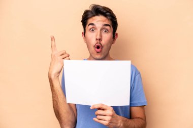 Beyazlı genç bir adam bej arka planda izole edilmiş bir pankart tutuyor. Harika bir fikri var, yaratıcılık kavramı..