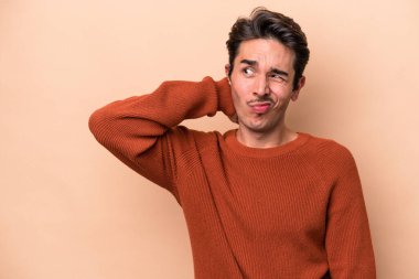 Kafkasyalı genç adam bej arka planda izole edilmiş kafasının arkasına dokunuyor, düşünüyor ve bir seçim yapıyor..