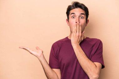 Beyaz tenli, bej arka planda izole edilmiş genç bir adam, avuç içi fotokopi alanı tutmaktan etkilenmiş..