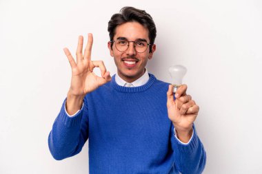 Beyaz arka planda izole edilmiş bir ampul tutan beyaz tenli genç adam neşeli ve kendinden emin bir hareket sergiliyor..