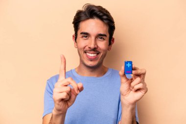 Beyazlı genç bir adam bej arka planda izole edilmiş bir bateri tutuyor. Bir numarayı parmağıyla gösteriyor..