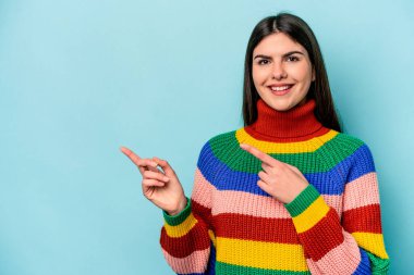 Mavi arka planda izole edilmiş genç beyaz kadın işaret parmağıyla işaret ederek heyecanlanıyor..