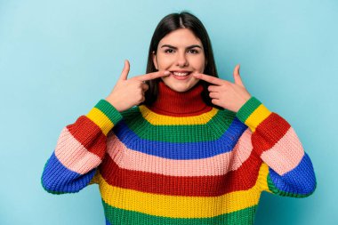 Mavi arka planda izole edilmiş genç beyaz kadın gülümsüyor, parmaklarını ağıza doğrultuyor..