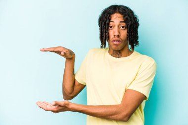 Genç Afrikalı Amerikalı bir adam mavi arka planda izole edilmiş. Elleri arasında bir kopya alanı tutarken şok olmuş ve hayrete düşmüştü..