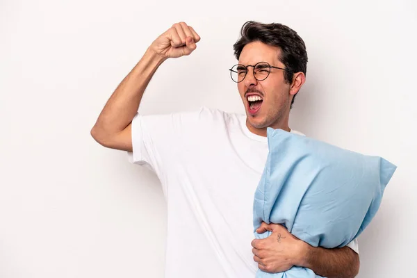 Young caucasian man wearing pajamas holding a pillow isolated on white background raising fist after a victory, winner concept.