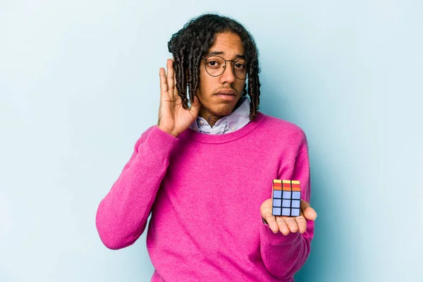 Young African American man holding a Rubiks cube isolated on blue background trying to listening a gossip.