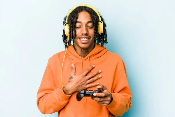 Young African American man playing with a video game controller isolated on blue background laughs out loudly keeping hand on chest.