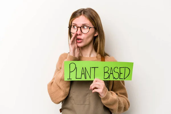 Young gardener woman holding a plan based placard isolated on white background is saying a secret hot braking news and looking aside