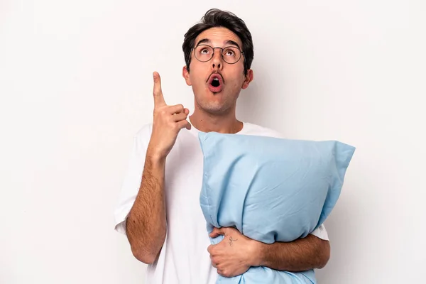 Young caucasian man wearing pajamas holding a pillow isolated on white background pointing upside with opened mouth.