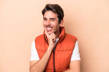 Beyaz tenli genç adam bej arka planda izole edilmiş mutlu ve kendinden emin gülümsüyor, elleriyle çenesine dokunuyor..