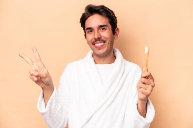 Elinde diş fırçası tutan beyaz bir bornoz giyen genç bir adam bej arka planda izole edilmiş neşeli ve kaygısız parmaklarıyla barış sembolü gösteriyor..