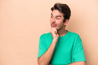 Beyaz tenli genç adam bej arka planda izole edilmiş. Rahatlamış bir şekilde fotokopi odasına bakan bir şey düşünüyor..