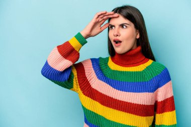 Kafkasyalı genç bir kadın mavi arka planda izole edilmiş, uzaklara bakıyor ve elini alnına koyuyor..