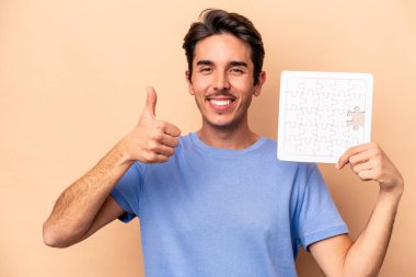 Beyazların arka planında izole edilmiş bir yapboz tutan genç beyaz adam gülümsüyor ve başparmağını kaldırıyor.