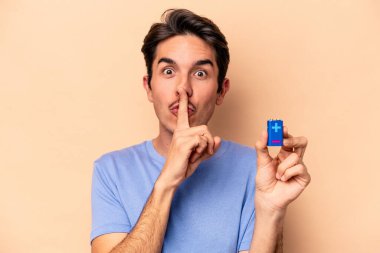 Beyazlı genç bir adam bej arka planda izole edilmiş bir bateriyi tutuyor ya sır saklıyor ya da sessizlik istiyor..