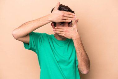 Beyazlı genç bir adam bej arka planda izole edilmiş. Kameraya parmaklarıyla göz kırpıyor, yüzünü kapatmaktan utanıyor..