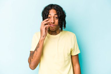 Genç Afrikalı Amerikalı bir adam mavi arka planda izole edilmiş ve parmakları dudaklarında bir sır saklıyor..