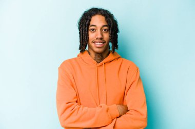 Genç Afro-Amerikalı, mavi arka planda izole edilmiş, kendinden emin, kararlılıkla kollarını kavuşturuyor..