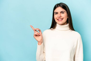 Mavi arka planda izole edilmiş genç beyaz kadın işaret parmağıyla işaret ederek gülümsüyor..