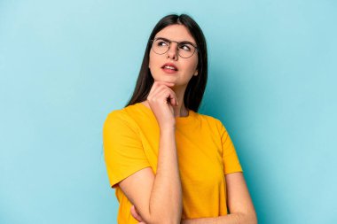 Kafkasyalı genç kadın mavi arka planda izole edilmiş düşünüyor ve yukarı bakıyor, yansıtıyor, düşünüyor, bir fantezi kuruyor..