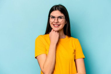 Mavi arka planda izole edilmiş genç beyaz kadın mutlu ve kendinden emin gülümsüyor, elleriyle çenesine dokunuyor..