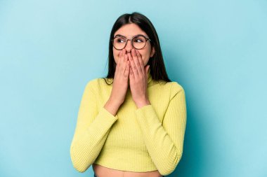 Mavi arka planda izole edilmiş genç beyaz kadın bir şeye gülüyor, elleriyle ağzını kapatıyor..