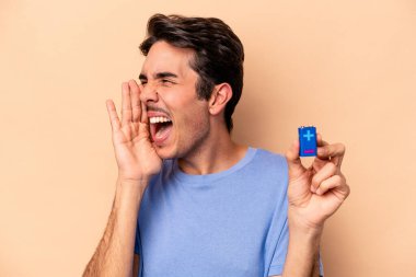 Beyazlı genç bir adam bej arka planda izole edilmiş bir bateriyi tutuyor bağırıyor ve avuç içini açık tutuyor..