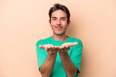 Beyaz tenli genç adam bej arka planda izole edilmiş. Elinde avuç içi olan bir şeyle kameraya sunuyor..